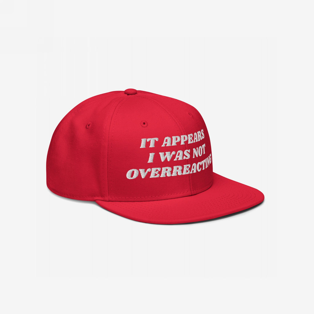 The Overreacting Hat is a bright red snapback cap with a flat brim, featuring a white embroidered slogan, IT APPEARS I WAS NOT OVERREACTING, symbolizing a stance against fascism and political gaslighting.