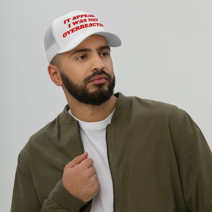 A young bearded man models the Overreacting Hat, a white mesh‐back trucker cap with bold red lettering, expressing defiance against political gaslighting while capturing a progressive spirit.