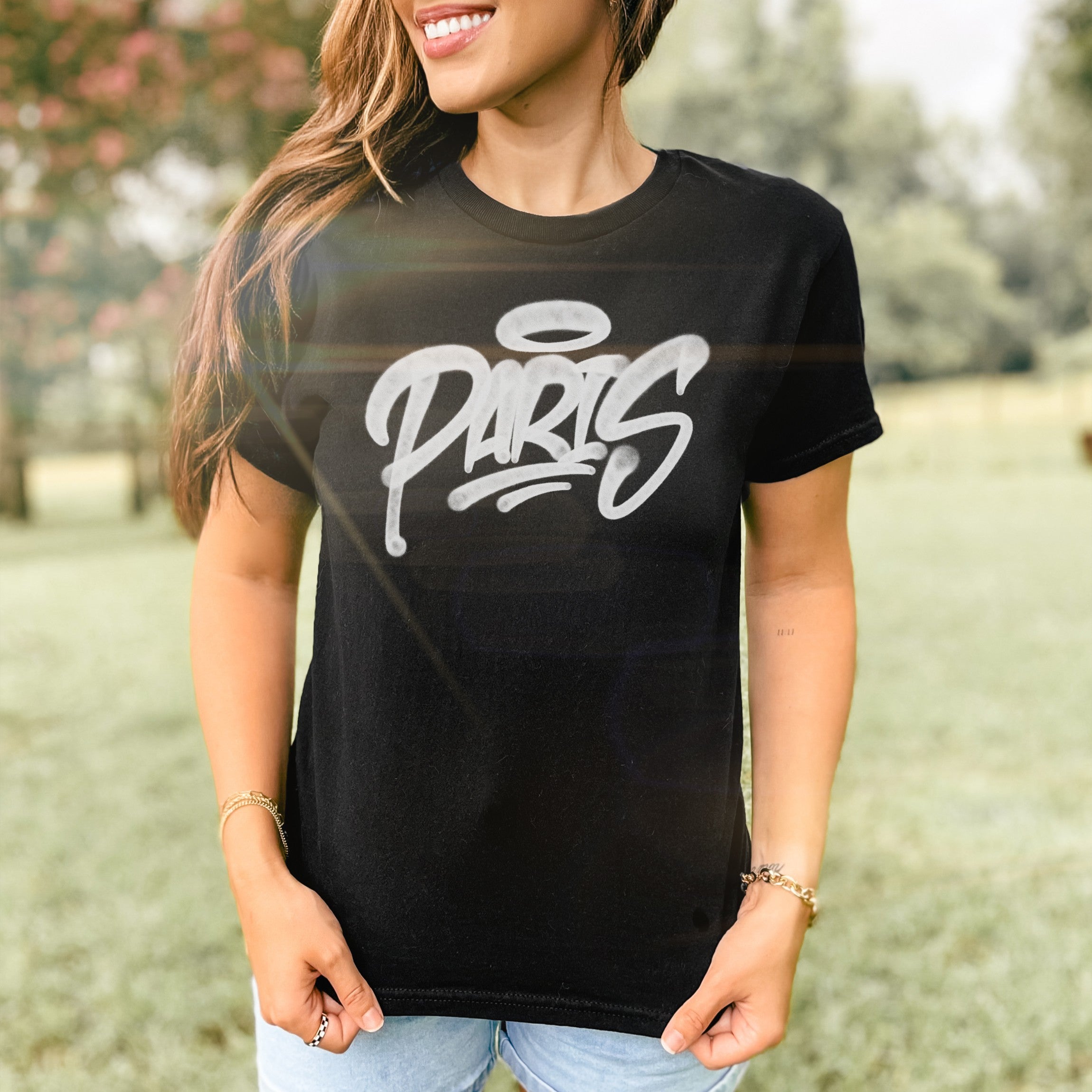 Woman outdoors wearing Paris Handstyle Women's Baby Tee: black with white graffiti PARIS and halo tag. Styled with light-wash jeans, bracelets, and rings, embodying bold, artistic expression.