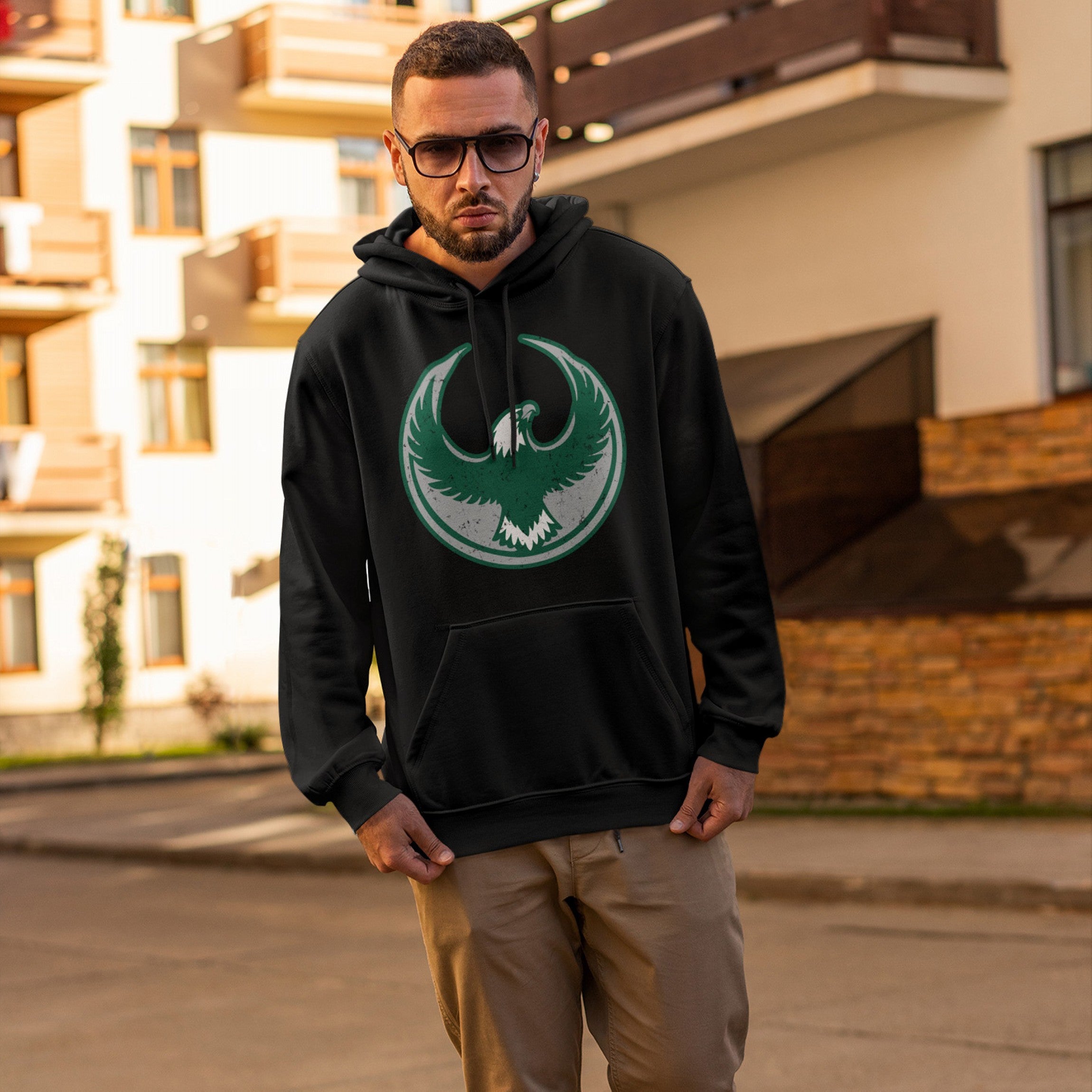 Young man wearing a Philly Rebel Alliance Hoodie, featuring a bold eagle emblem, stands in front of a modern building. The hoodie embodies anti-fascist sentiments, supporting equality and immigrant rights.