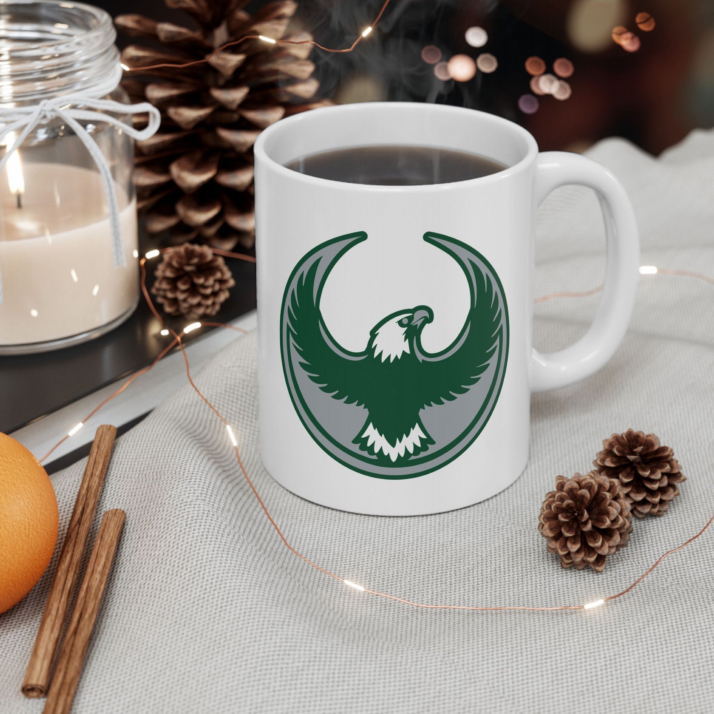 Philly Rebel Alliance Mug with a stylized eagle emblem, filled with steaming coffee on a holiday-themed tabletop, surrounded by pine cones, cinnamon sticks, a tangerine, and a jar candle.