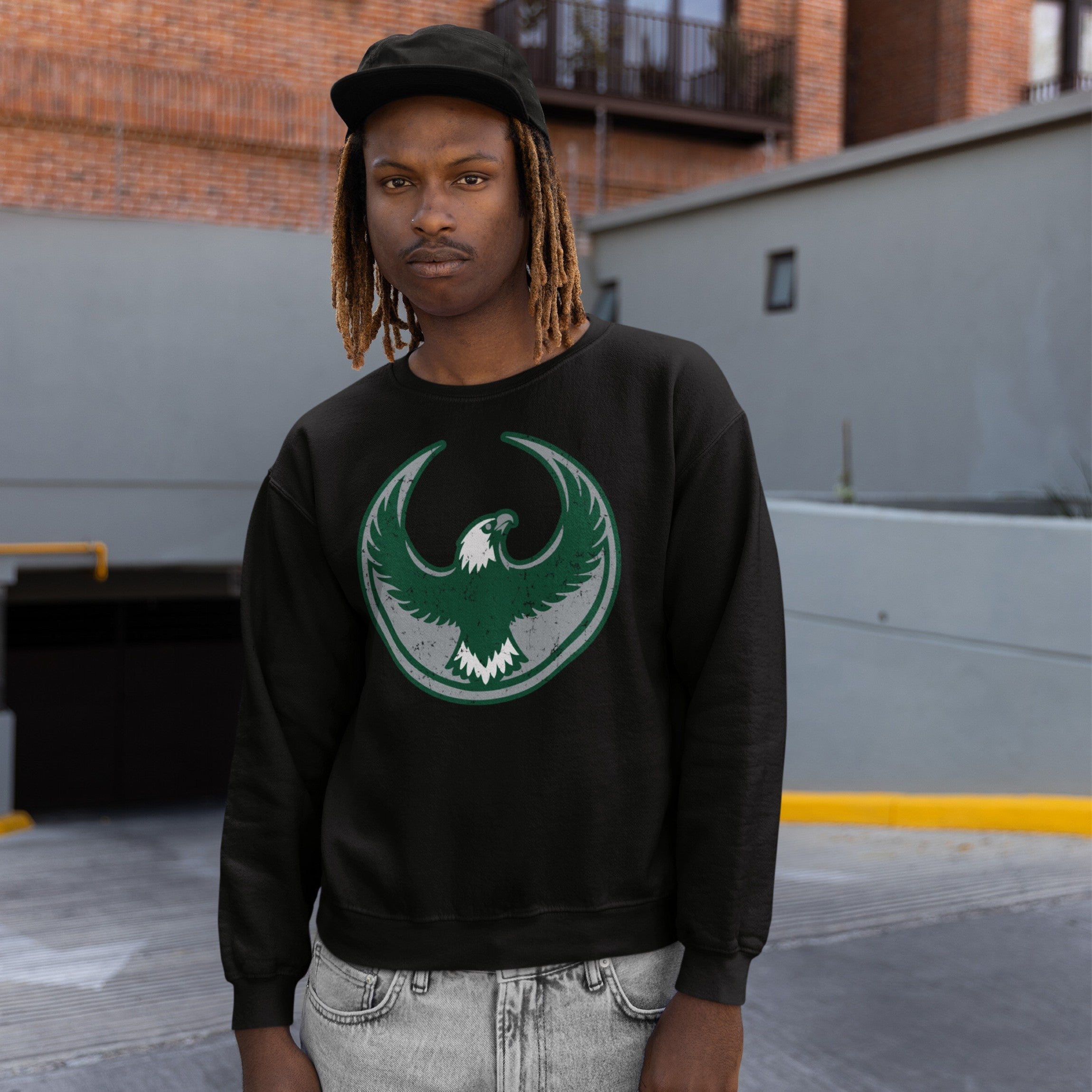 Young man in urban setting wears a Philly Rebel Alliance Hoodie, featuring a green-and-white eagle emblem, embodying anti-fascist and pro-equality values. He's styled in a black crewneck with light gray jeans.
