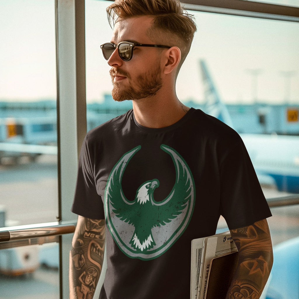 Man at airport wearing a Philly Rebel Alliance T-shirt with a bold eagle emblem, symbolizing anti-fascist protest. He's holding a newspaper, showcasing the shirt's message of standing for immigrant rights.