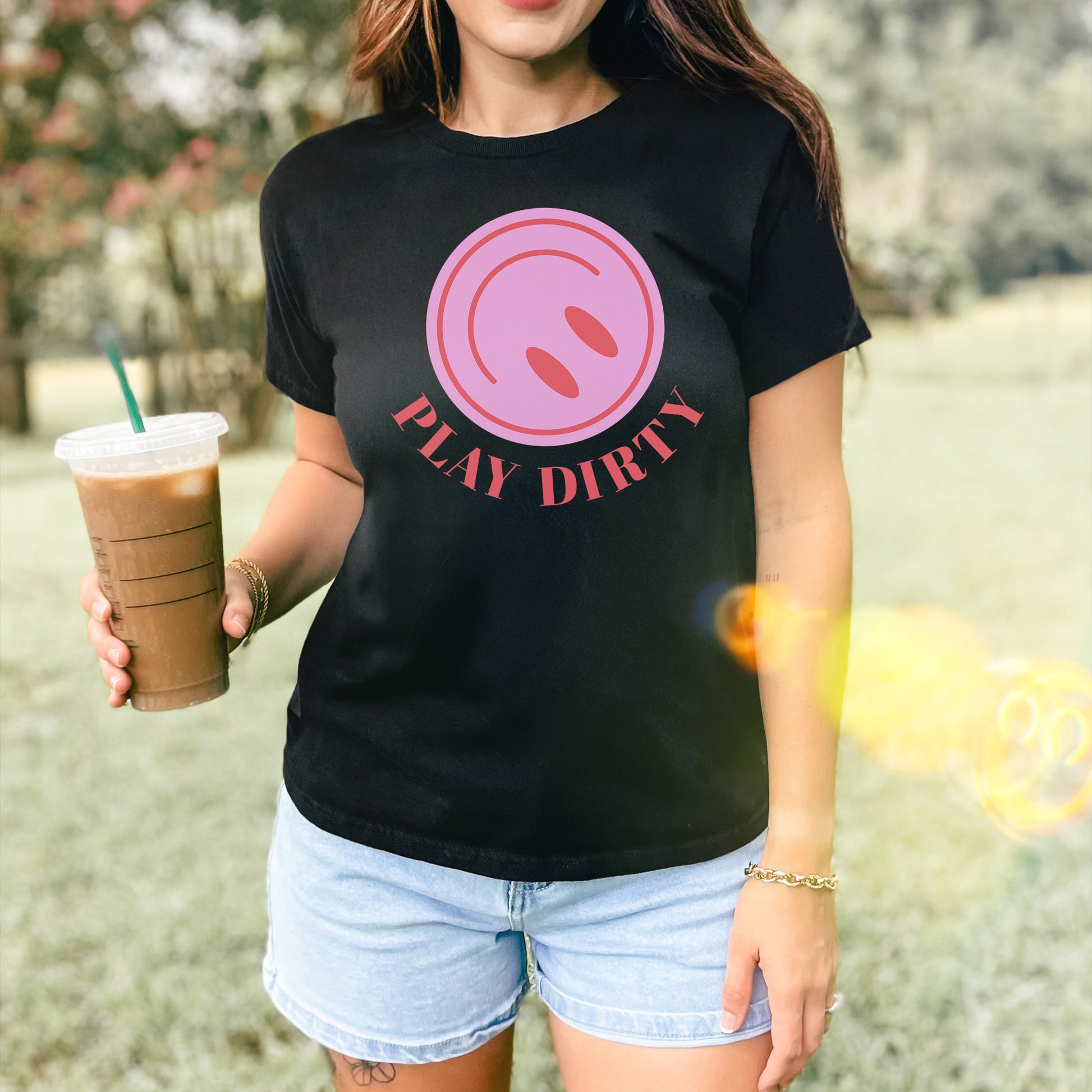 Person wearing a Play Dirty Women's Baby Tee with a pink circular graphic, holding iced coffee, and sporting a gold chain bracelet, embodying a bold, flirty style perfect for adventurous outings.