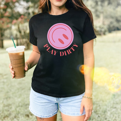 Person wearing a Play Dirty Women's Baby Tee with a pink circular graphic, holding iced coffee, and sporting a gold chain bracelet, embodying a bold, flirty style perfect for adventurous outings.