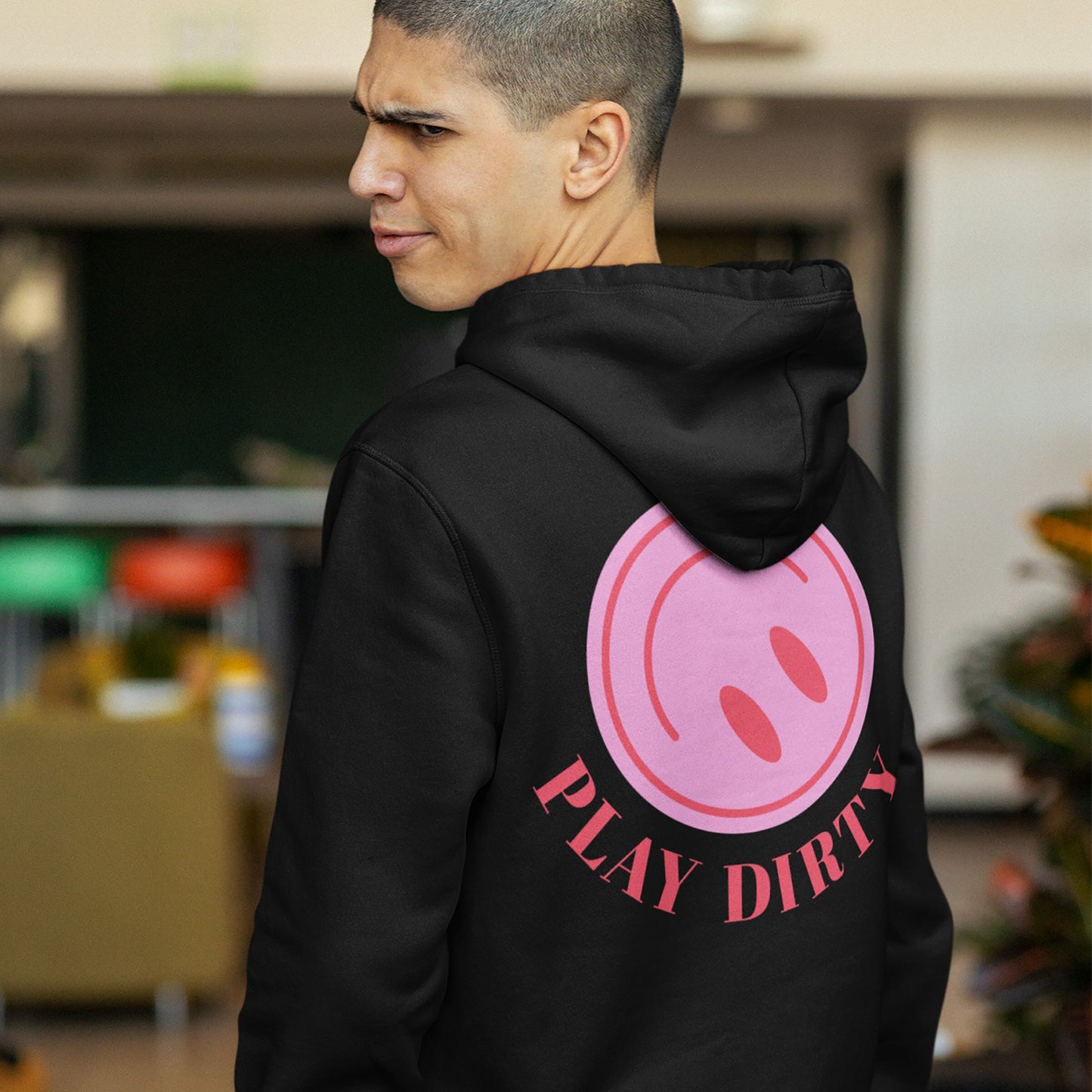 Young man in a black Play Dirty hoodie featuring a large pink smiley face and bold PLAY DIRTY text, embodying a rebellious style. Image taken in a casual indoor setting.