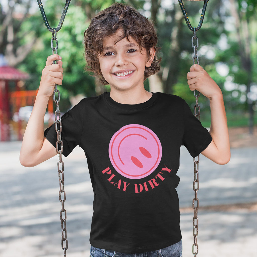 Young boy smiling on a swing, wearing the Play Dirty T-shirt featuring a pink circular graphic and bold red text, embodying playful chaos and carefree fun.