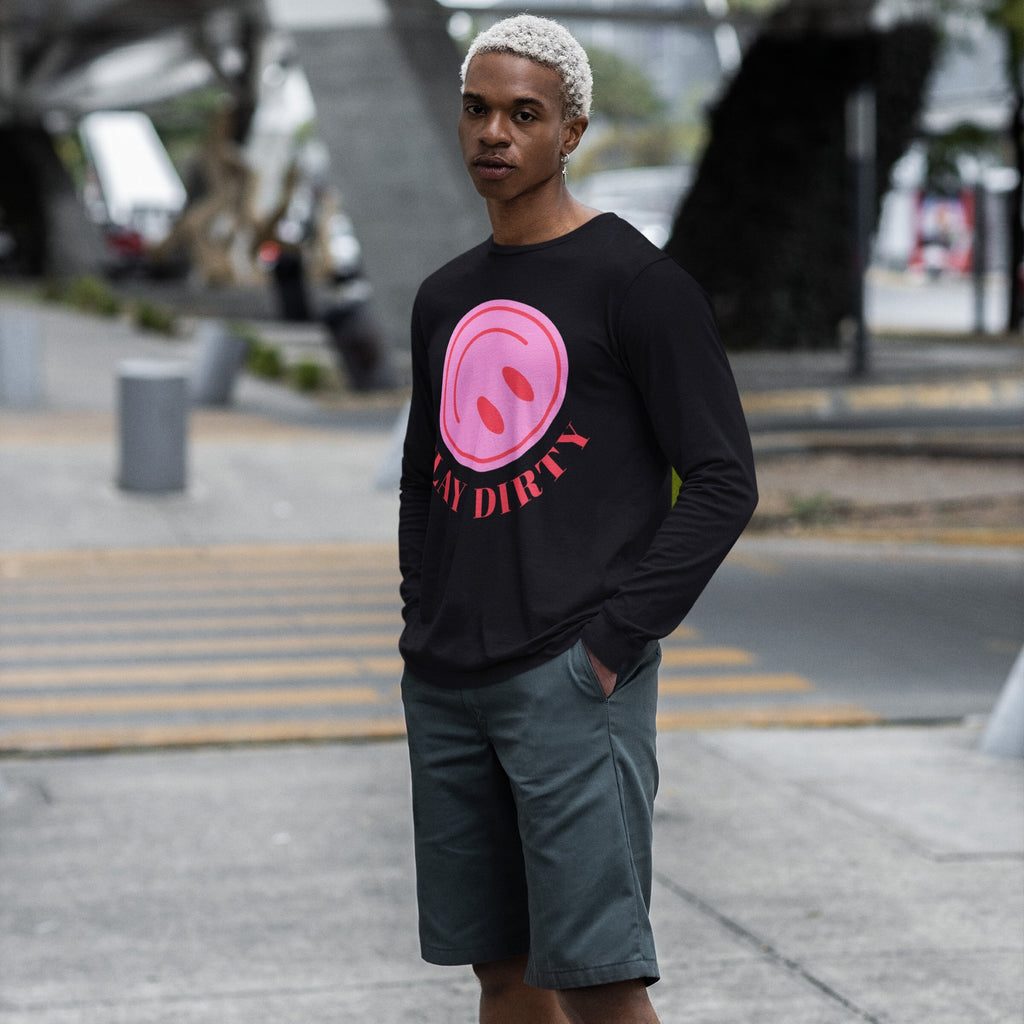 Young man wearing a Play Dirty T-shirt with a pink smiling-face graphic and “PLAY DIRTY” text, paired with dark-green shorts, standing on a street.