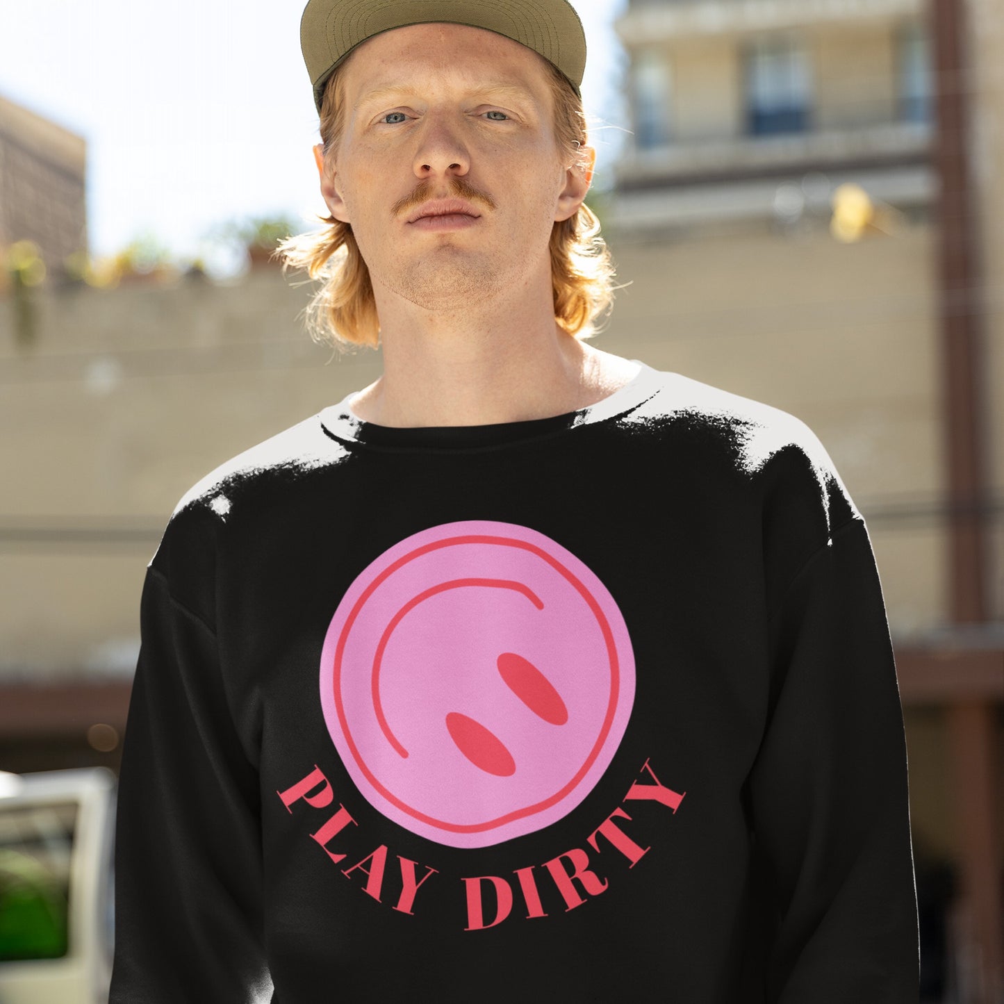 Young man in a “Play Dirty” sweatshirt with a pink graphic and bold red slogan, embodying mischief and subversion, set against an urban backdrop.