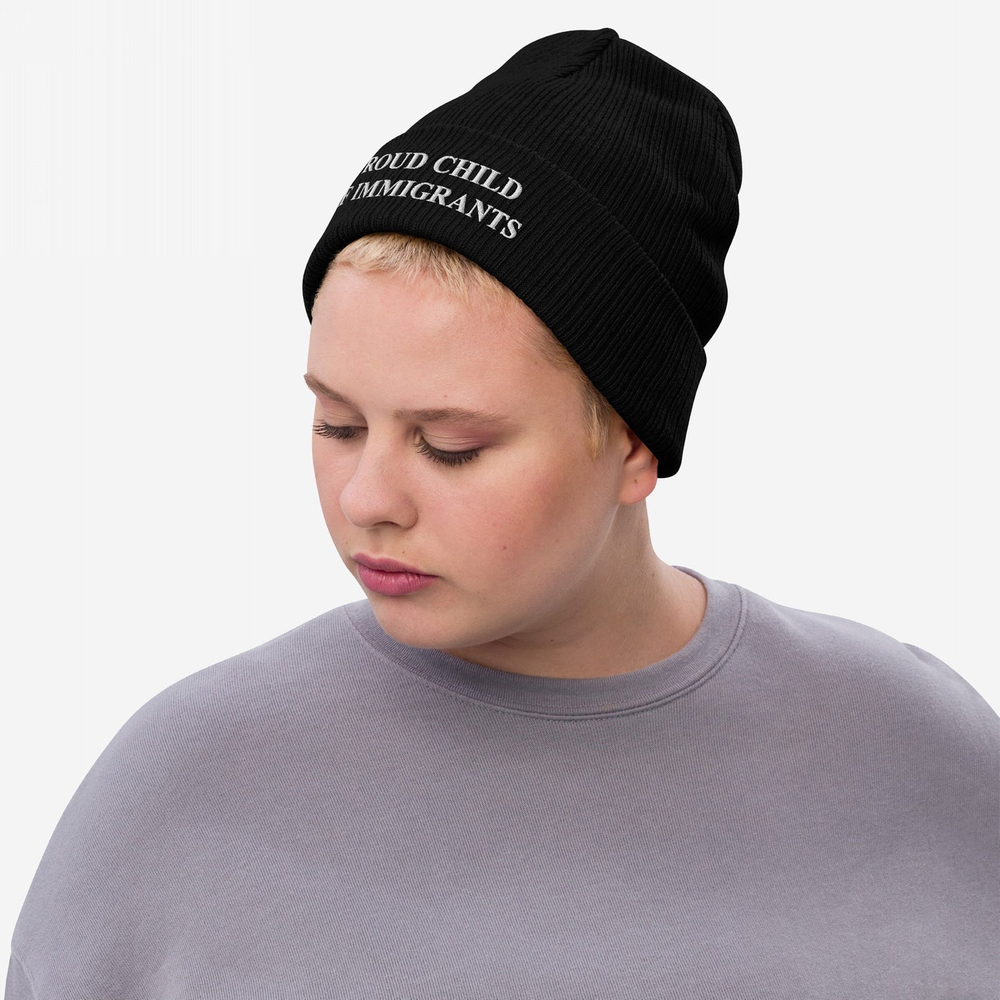Young person with short blonde hair wears the Proud Child Of Immigrants Hat, a black ribbed beanie with bold embroidery, symbolizing identity and resilience. Their light gray top complements the beanie’s statement.
