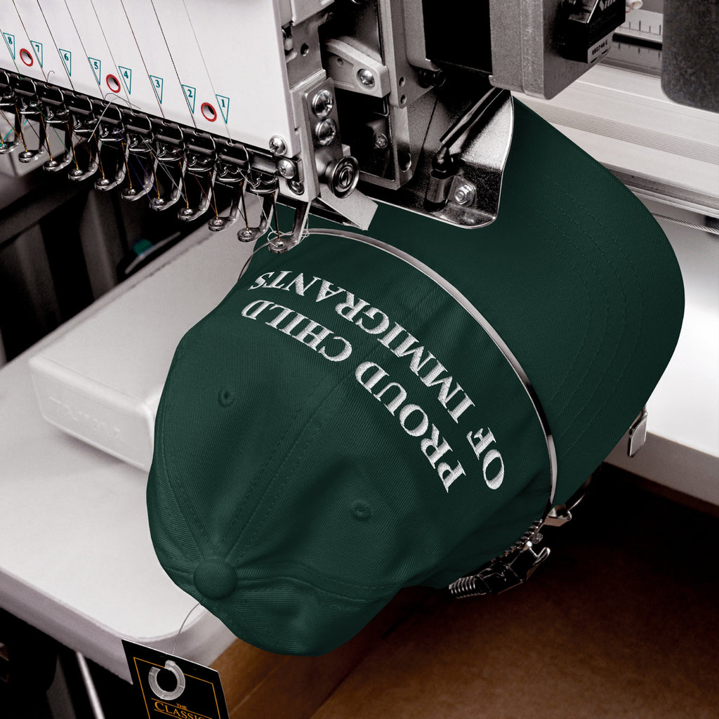 Green cap in embroidery machine, displaying PROUD CHILD OF IMMIGRANTS in white thread, symbolizing resilience and pride.