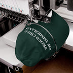 Green cap in embroidery machine, displaying PROUD CHILD OF IMMIGRANTS in white thread, symbolizing resilience and pride.