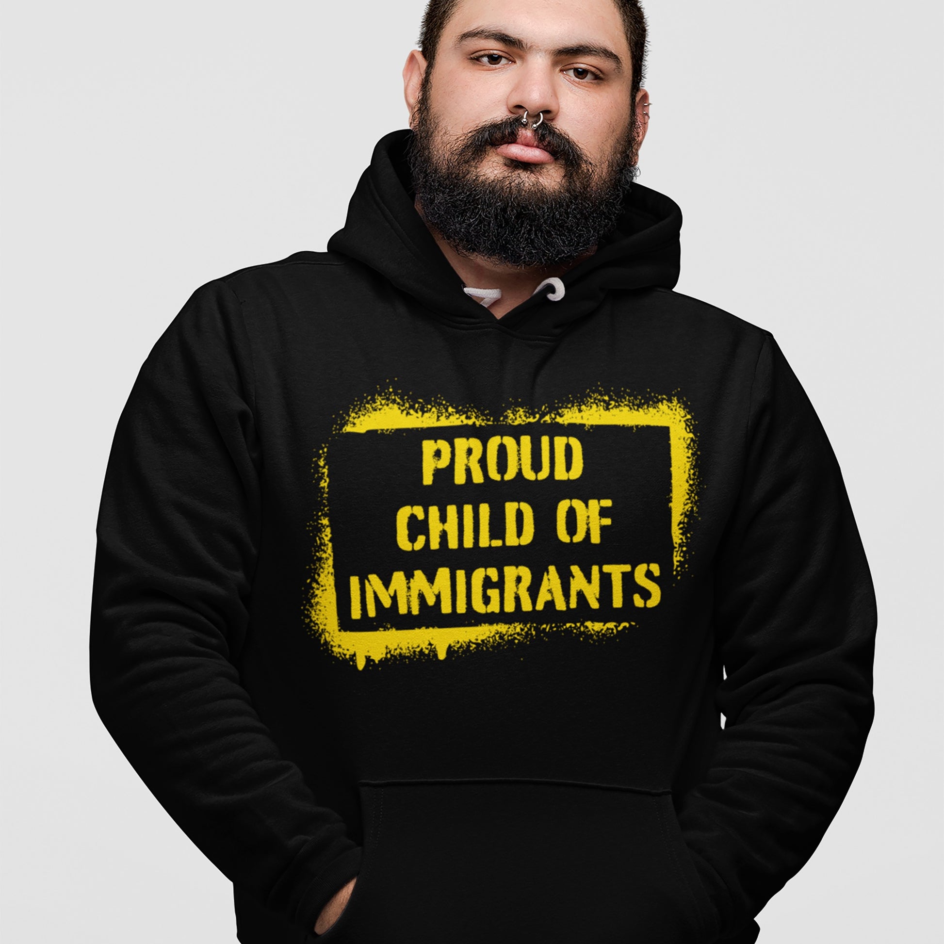 Proud Child Of Immigrants Hoodie features a spray-painted yellow frame with bold text, worn by a person with a dark beard and septum piercing, embodying heritage and identity.