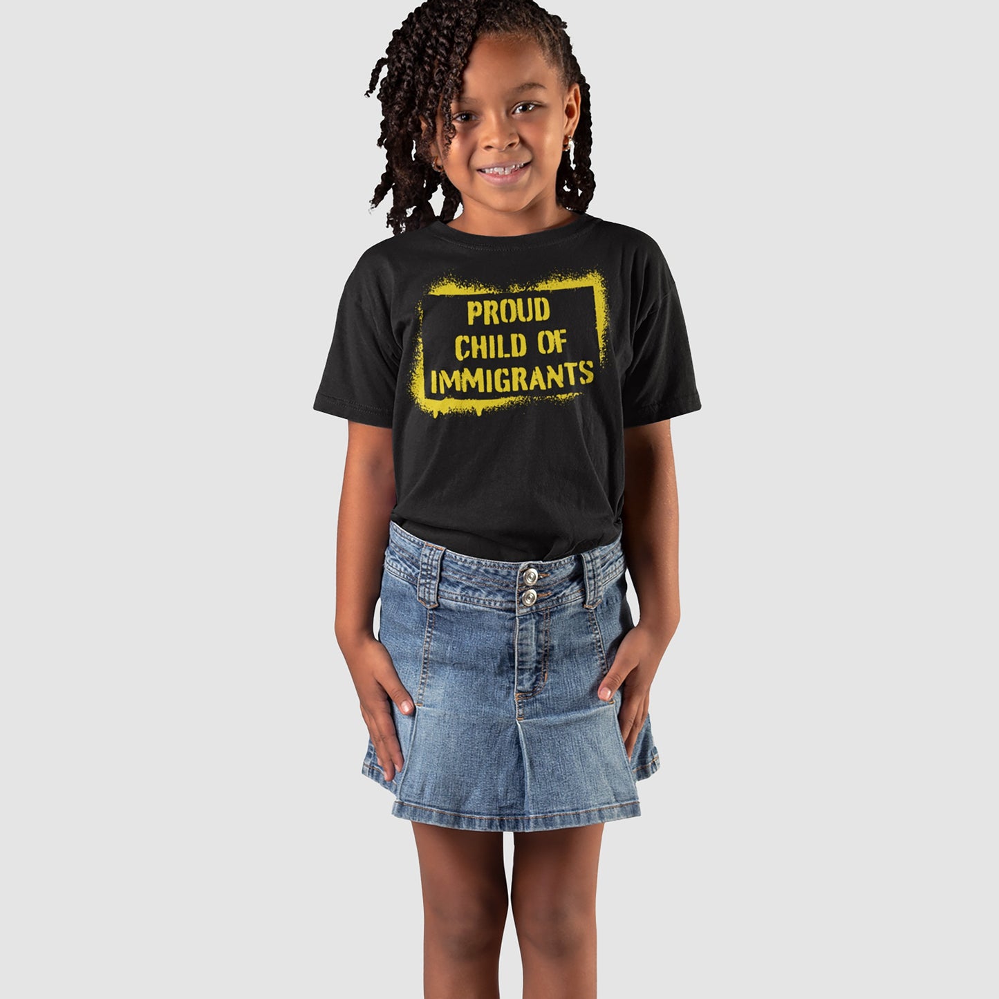 A young girl wears the Proud Child of Immigrants T-shirt, featuring bold yellow text, paired with a blue denim skirt, embodying cultural pride and the legacy of migration.