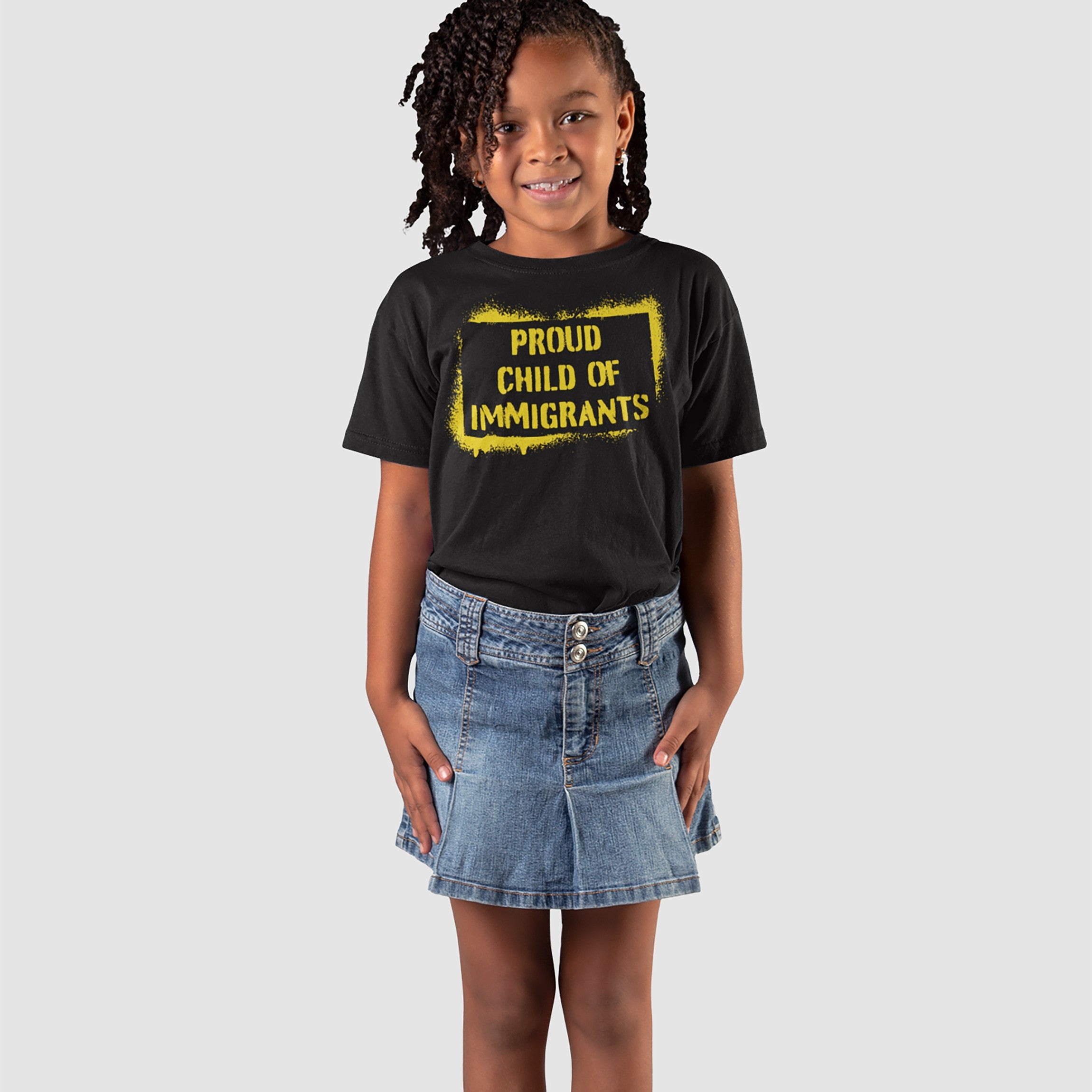 A young girl wears the Proud Child of Immigrants T-shirt, featuring bold yellow text, paired with a blue denim skirt, embodying cultural pride and the legacy of migration.