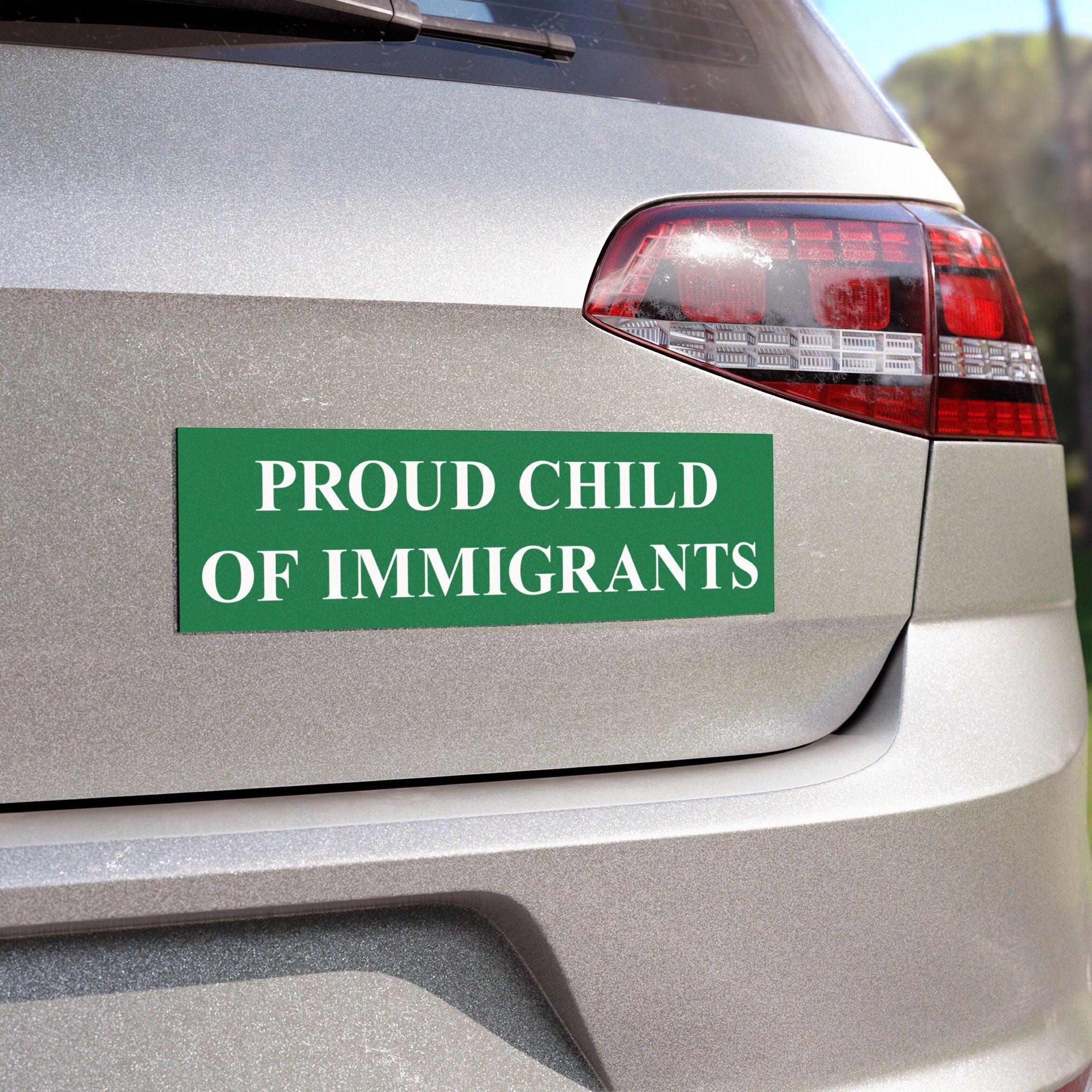Proud Child Of Immigrants Sticker & Magnet displayed on a silver car's rear, featuring bold white text on a green background. Part of the taillight and blurred foliage are visible.
