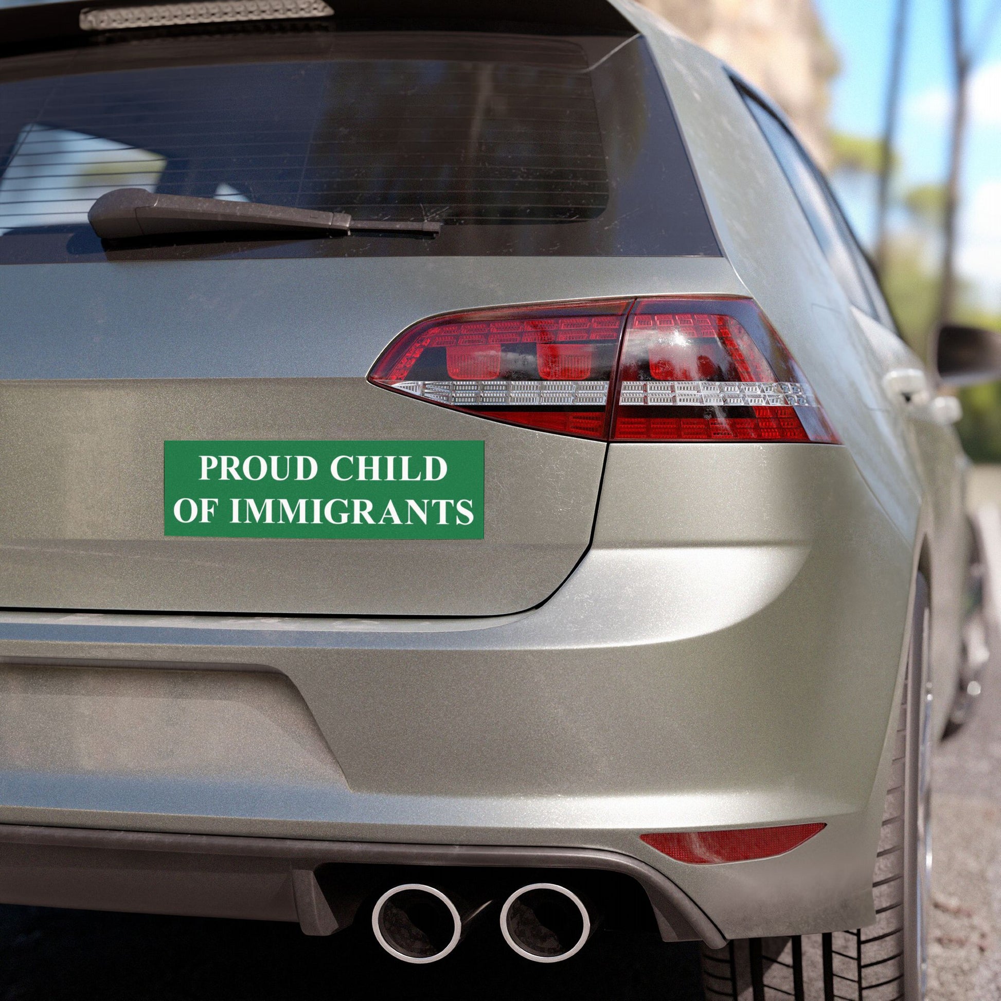 The rear of a silver-gray hatchback displays a Proud Child Of Immigrants Sticker & Magnet on its tailgate, showcasing a green backdrop with white text, celebrating immigrant heritage and resilience.
