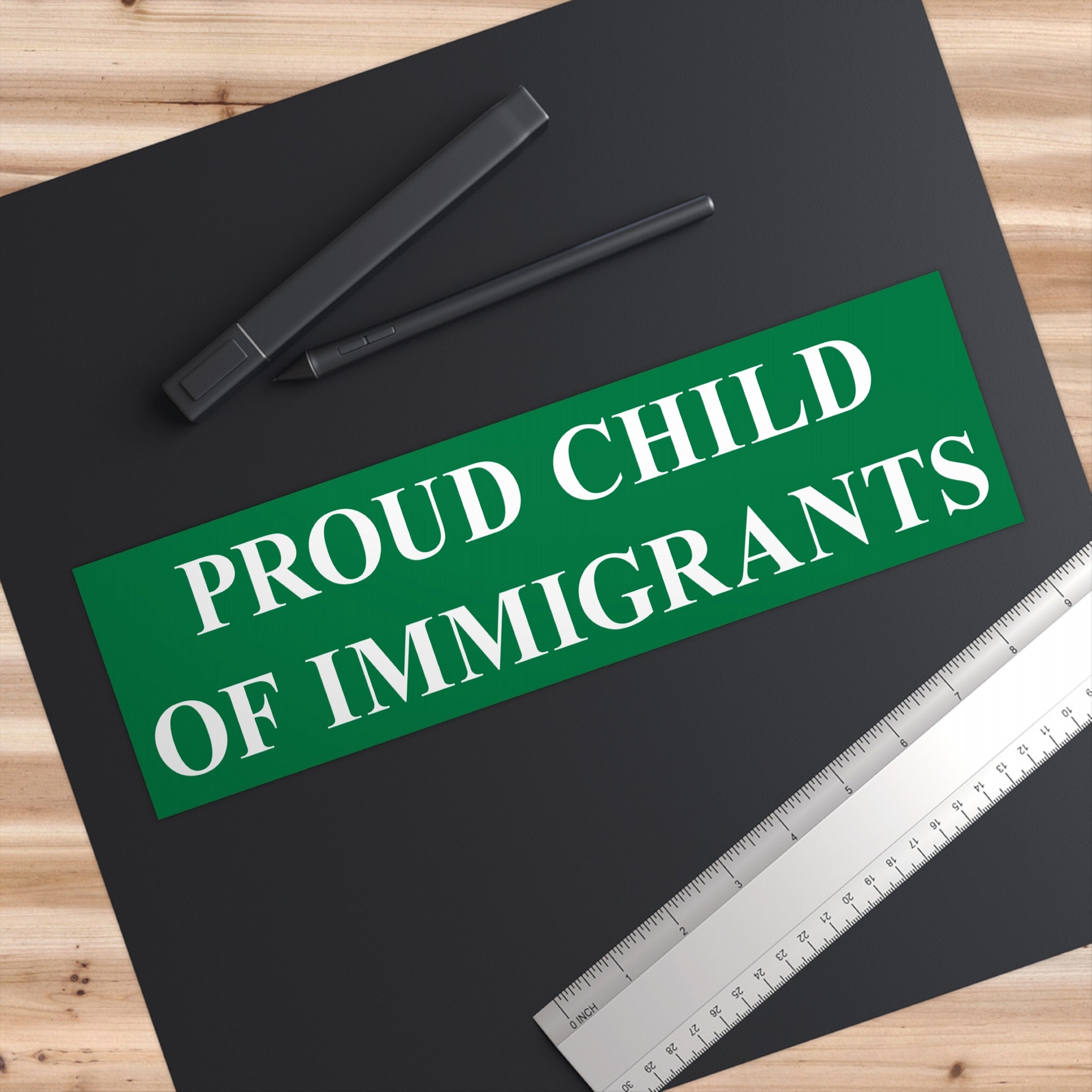 Proud Child Of Immigrants Sticker & Magnet on a black desk mat with a digital stylus and metal ruler, celebrating heritage and resilience against anti-immigrant sentiment with bold, empowering white text.