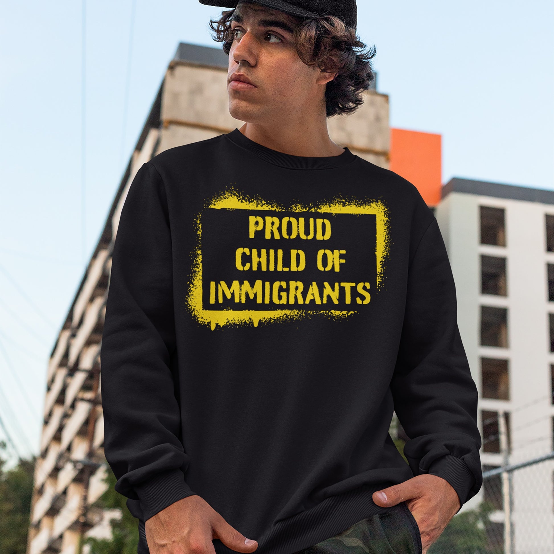 Young man wearing the Proud Child Of Immigrants crewneck sweatshirt with bold yellow text, symbolizing heritage and migration stories, stands against an urban backdrop.
