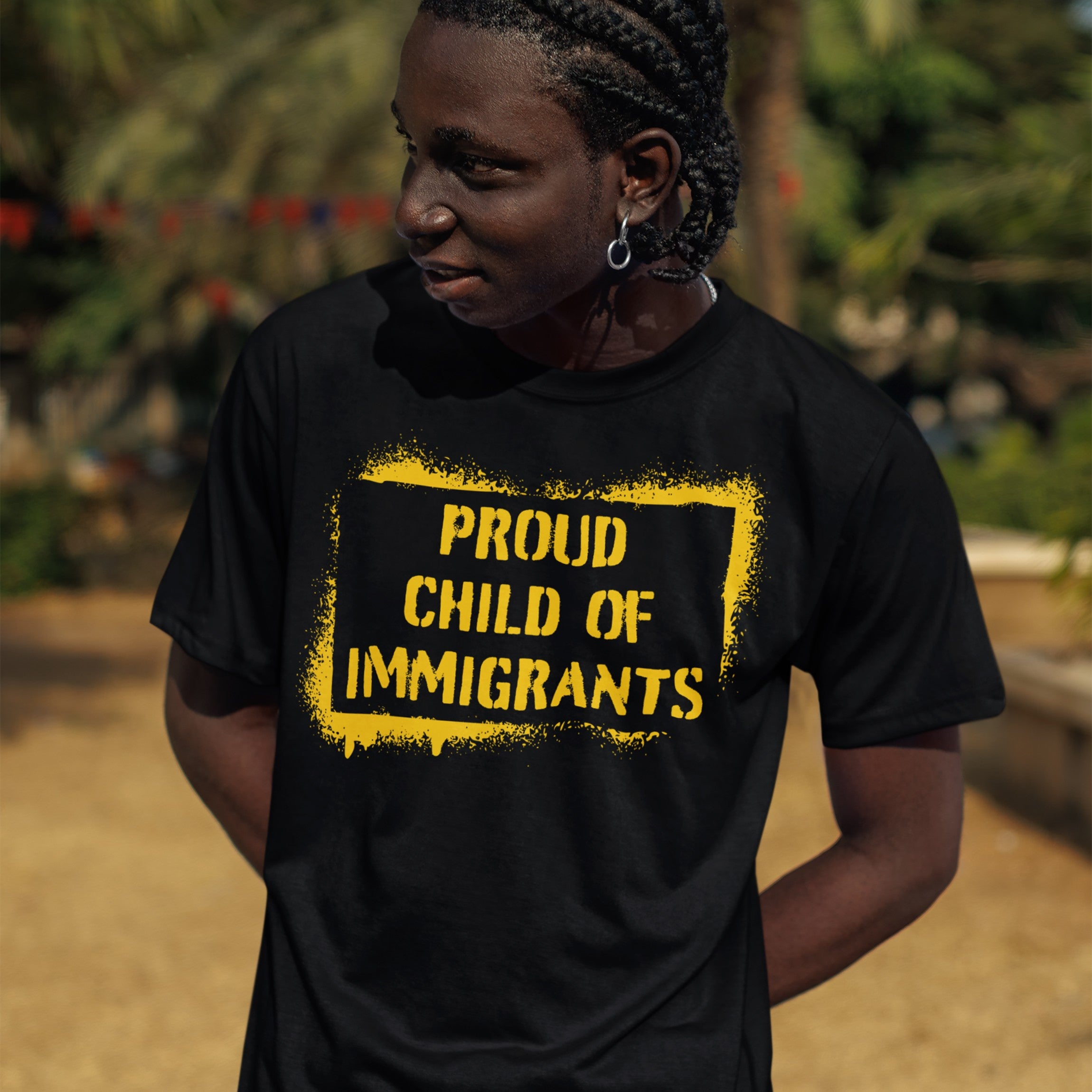 Young person outdoors wearing a Proud Child of Immigrants T-shirt, featuring bold yellow lettering, representing cultural pride and strength. The shirt embodies belonging and shared stories across generations.