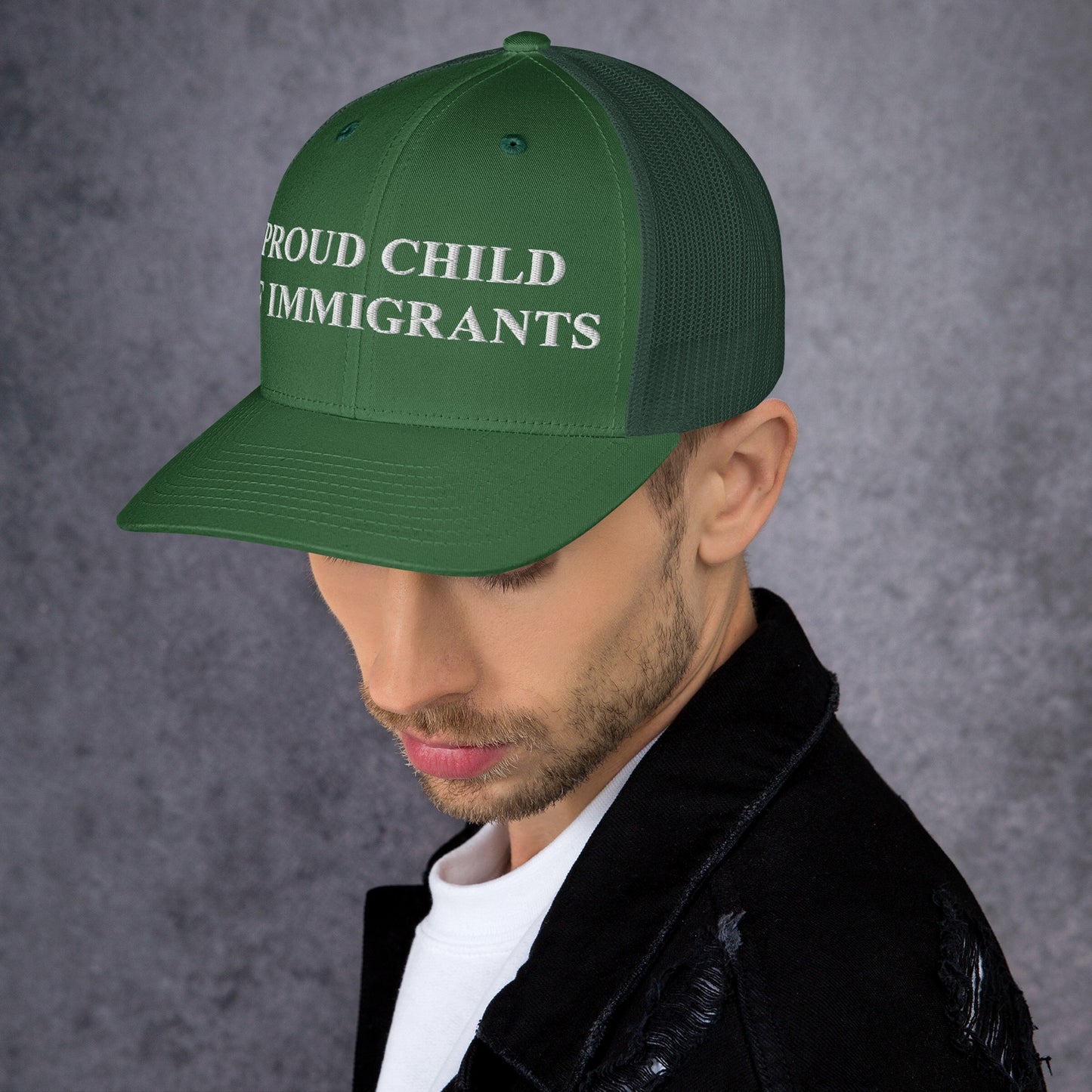 Young man wearing a Proud Child of Immigrants Hat, a green mesh-back cap with bold white embroidered text, symbolizing resilience and identity. The hat complements his black jacket and white shirt ensemble.