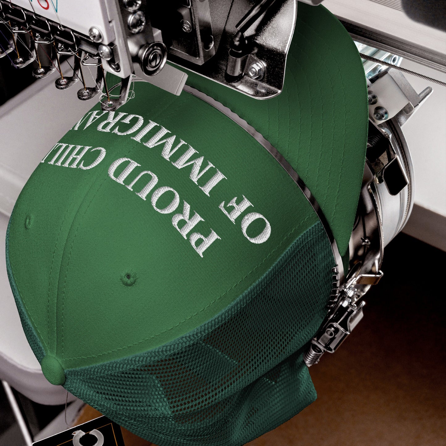 Green trucker-style baseball cap in embroidery machine, showcasing PROUD CHILD OF IMMIGRANTS text being stitched, embodying resilience and pride, ideal for those honoring roots and identity.