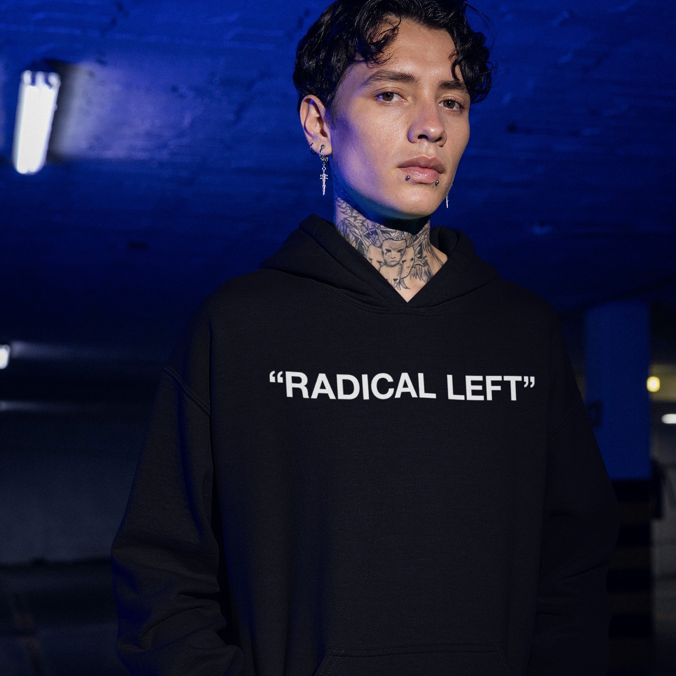 Young person wearing the Radical Left hoodie in a moody, urban setting. The black hoodie features bold white text, embodying activism and defiance, in line with supporting equality and human rights.