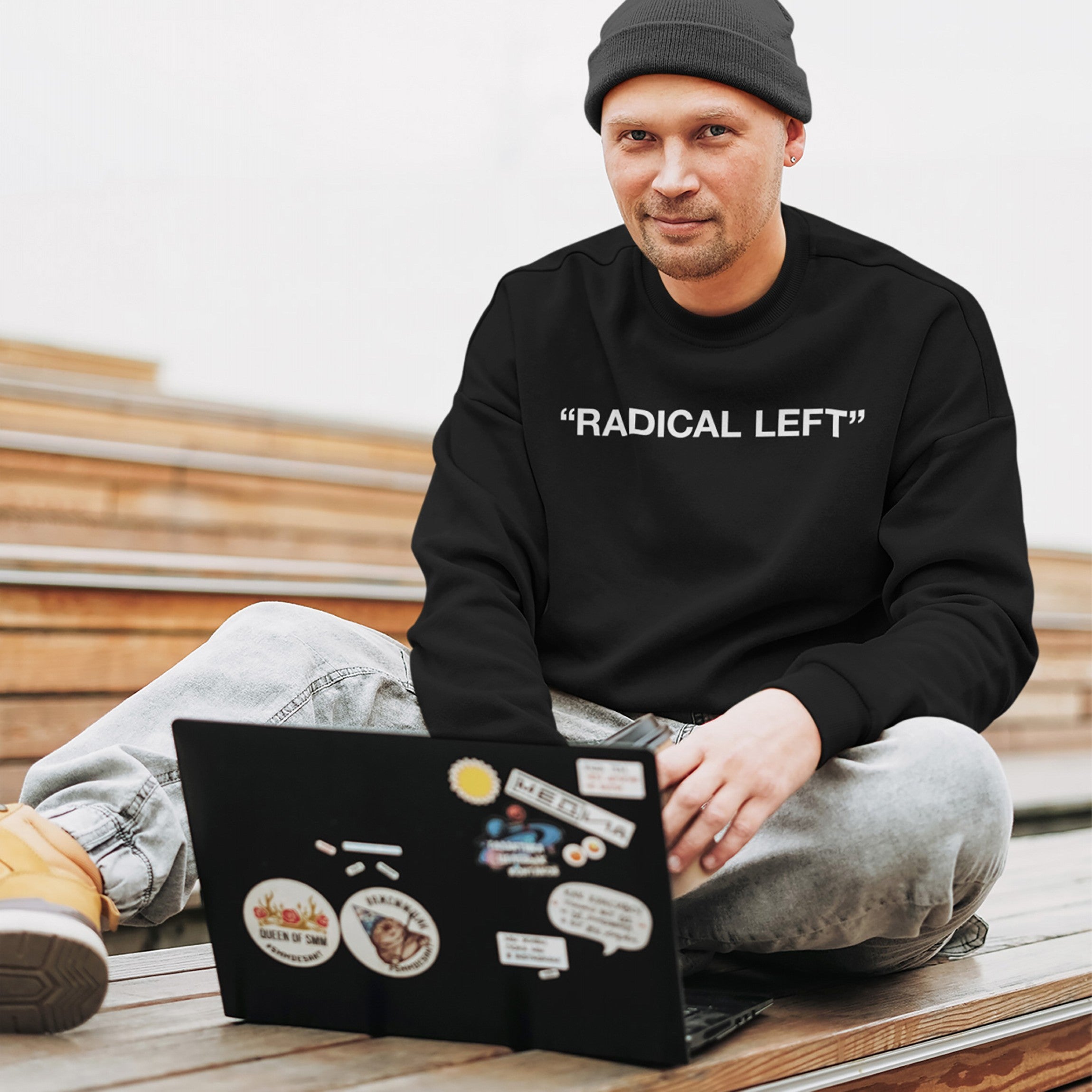 Man in light-wash jeans and boots wears Radical Left sweatshirt, embodying activism and equality while working on a sticker-covered laptop, reflecting the store's commitment to social justice and empowerment.