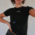 Snacks Women's Baby Tee on a person, showcasing the bold SNACKS print. The cropped, Y2K-style design complements dark jeans, embodying quirky, unapologetic vibes with rainbow light reflections enhancing its playful flair.