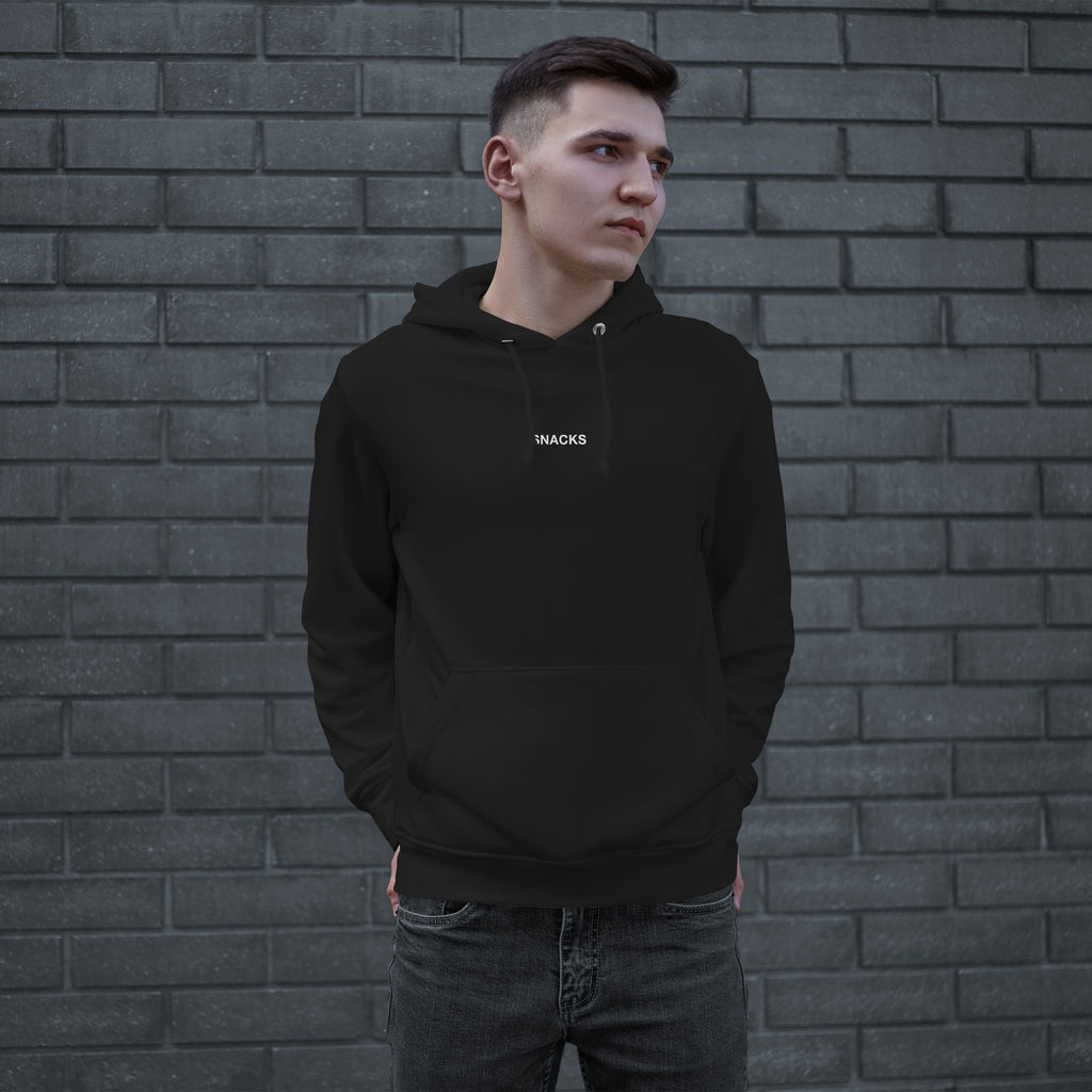 Young man in front of a brick wall wearing the Snacks Hoodie, a black pullover with SNACKS in small white letters, embodying a minimalist style that appreciates simple pleasures.