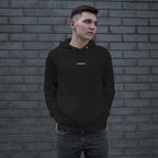 Young man in front of a brick wall wearing the Snacks Hoodie, a black pullover with SNACKS in small white letters, embodying a minimalist style that appreciates simple pleasures.
