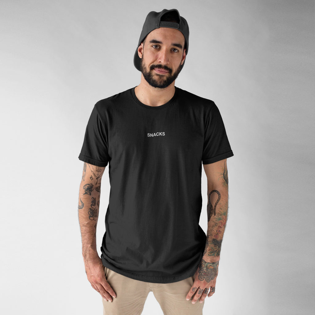 Young man models the Snacks T-shirt, featuring “SNACKS” in white on black. He sports light pants, a backward cap, and tattoos, embodying the playful, edgy spirit of this understated tee.