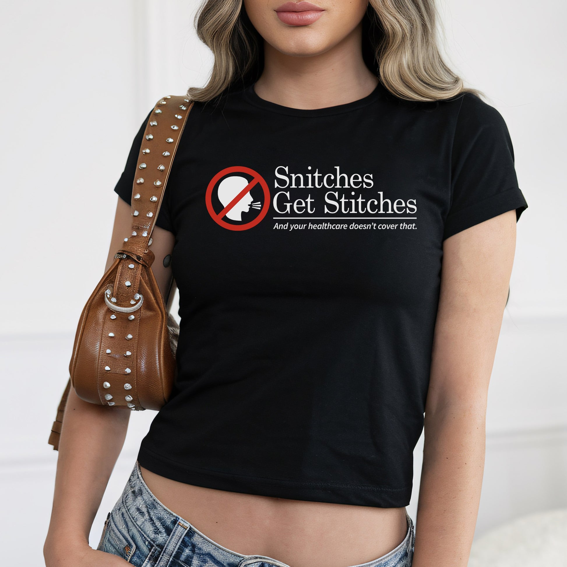 Young woman in a fitted Snitches Get Stitches Women's Baby Tee with bold text critiquing healthcare, paired with light-wash jeans and a studded leather shoulder bag, embodying edgy, socially conscious style.