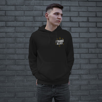 Young man wearing Social Club Hoodie featuring STRANGE ALLIES graphic on chest, standing against a gray brick wall, hands in pockets, embodying a blend of humor and supportive vibes.