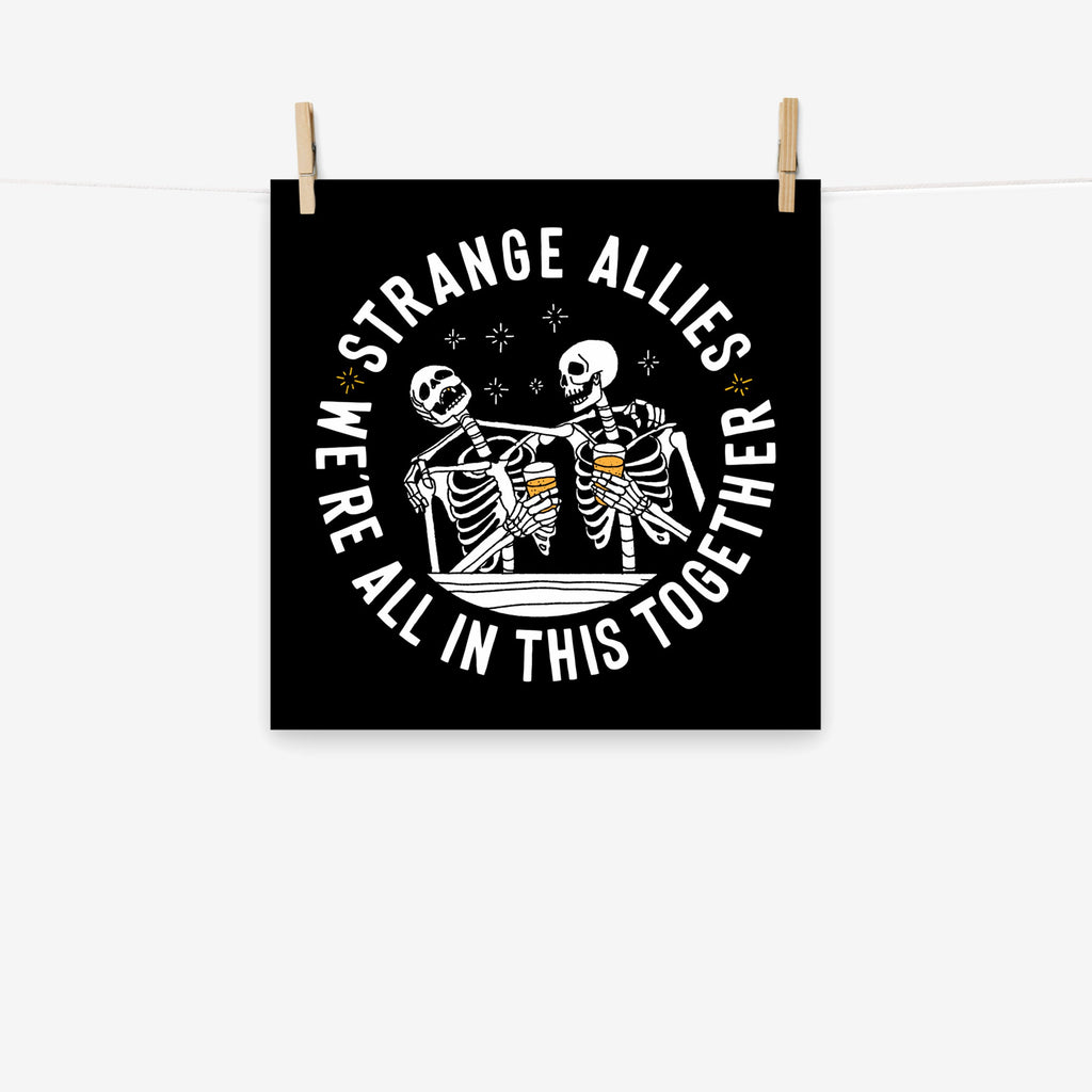 Social Club Giclée Print features two cartoon-style skeletons with beer glasses, surrounded by sparkles and the bold message, “WE’RE ALL IN THIS TOGETHER” on a square black background.
