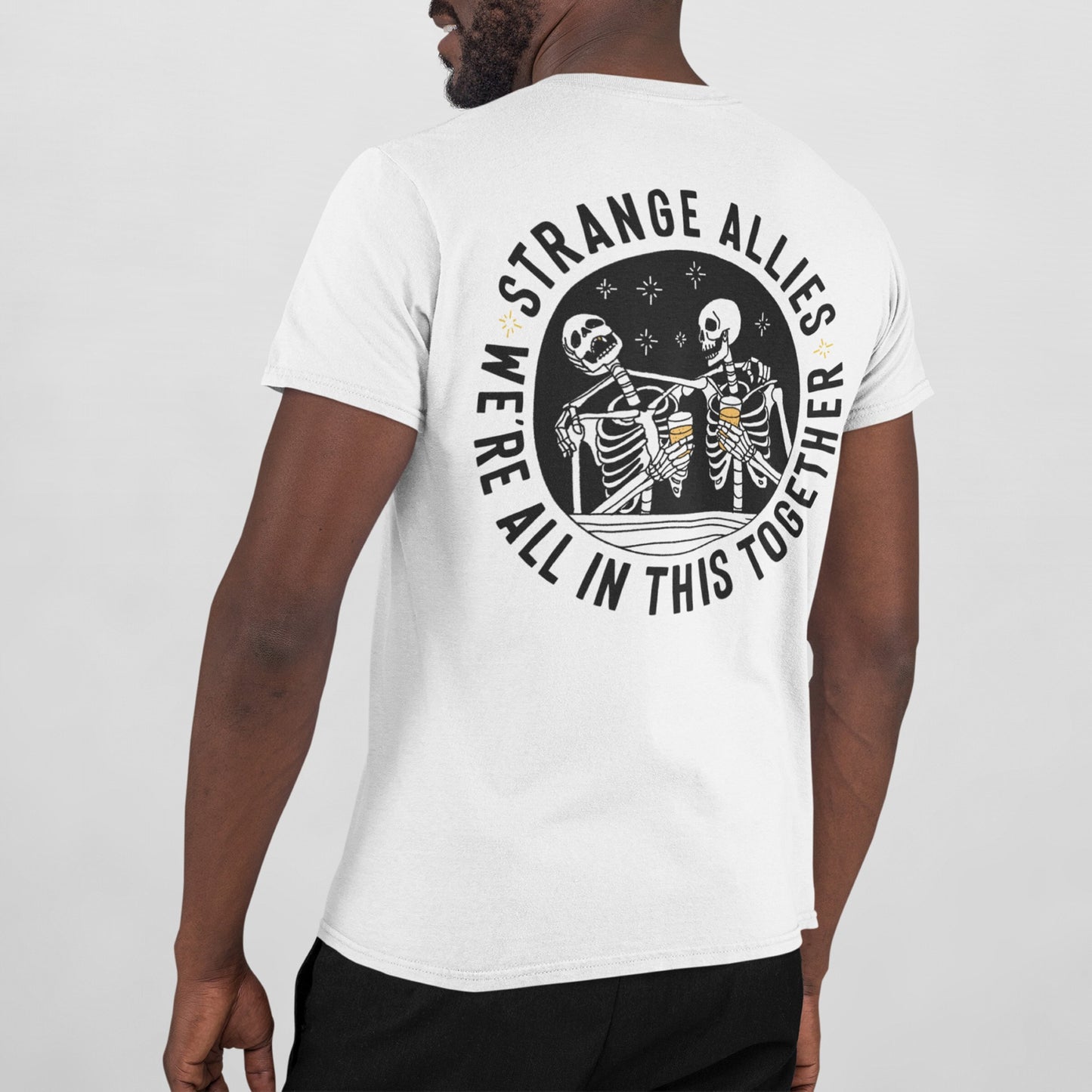 The Social Club T-shirt's back displays skeletons clinking beer mugs under stars, encircled by “STRANGE ALLIES” and “WE’RE ALL IN THIS TOGETHER,” capturing camaraderie among unexpected friends.
