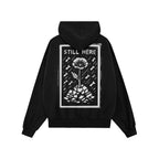 Still Here JULZ Hoodie featuring a bold graphic of a resilient flower growing through rubble amid falling shapes, symbolizing