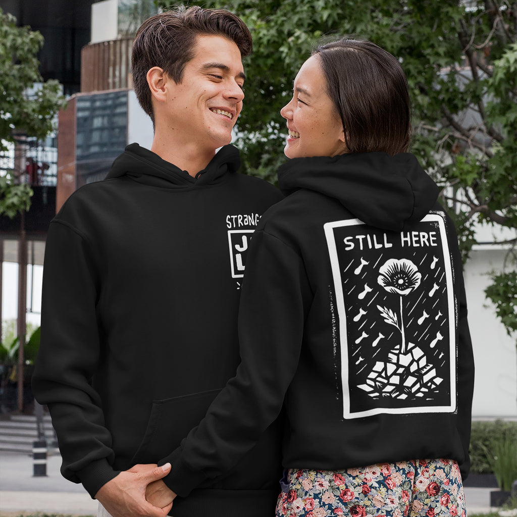Young couple outdoors, both in black hooded Still Here JULZ Hoodies, featuring protest art with a flower on rubble. The man’s hoodie has a small front logo, woman’s displays art on the back.