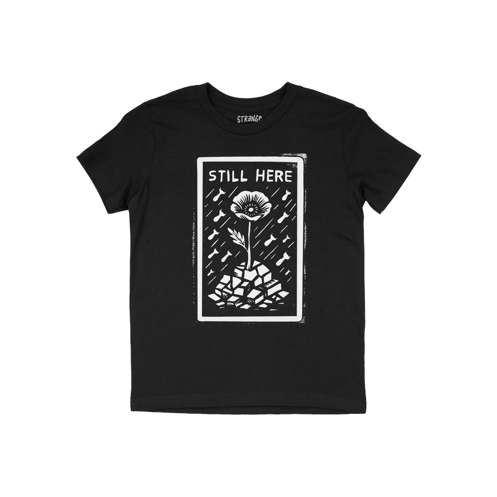 Still Here T-shirt features a bold white graphic of a flower emerging from rubble with bombs falling, symbolizing resilience 