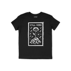 Still Here T-shirt features a bold white graphic of a flower emerging from rubble with bombs falling, symbolizing resilience 