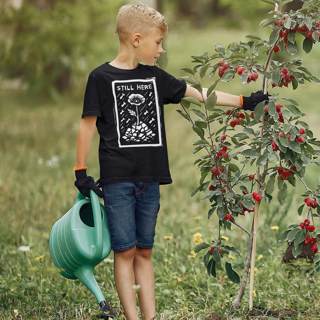 A young boy wearing a STILL HERE T-shirt inspects a fruit tree in a garden, symbolizing resilience and protest. The shirt features a flower-and-rubble design, aligning with anti-war and human rights messages.