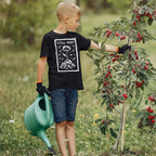A young boy wearing a STILL HERE T-shirt inspects a fruit tree in a garden, symbolizing resilience and protest. The shirt features a flower-and-rubble design, aligning with anti-war and human rights messages.
