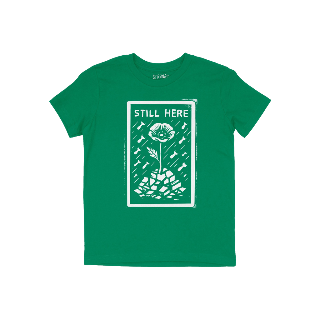 Still Here T-shirt features a white graphic of a flower emerging from rubble amid falling bombs, symbolizing resilience and p