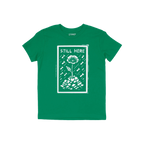 Still Here T-shirt features a white graphic of a flower emerging from rubble amid falling bombs, symbolizing resilience and p