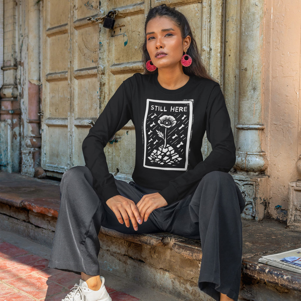 Person wearing the Still Here T-shirt with a flower graphic symbolizing resilience amidst destruction, seated by a weathered wooden door, conveying a powerful anti-war and pro-freedom message.