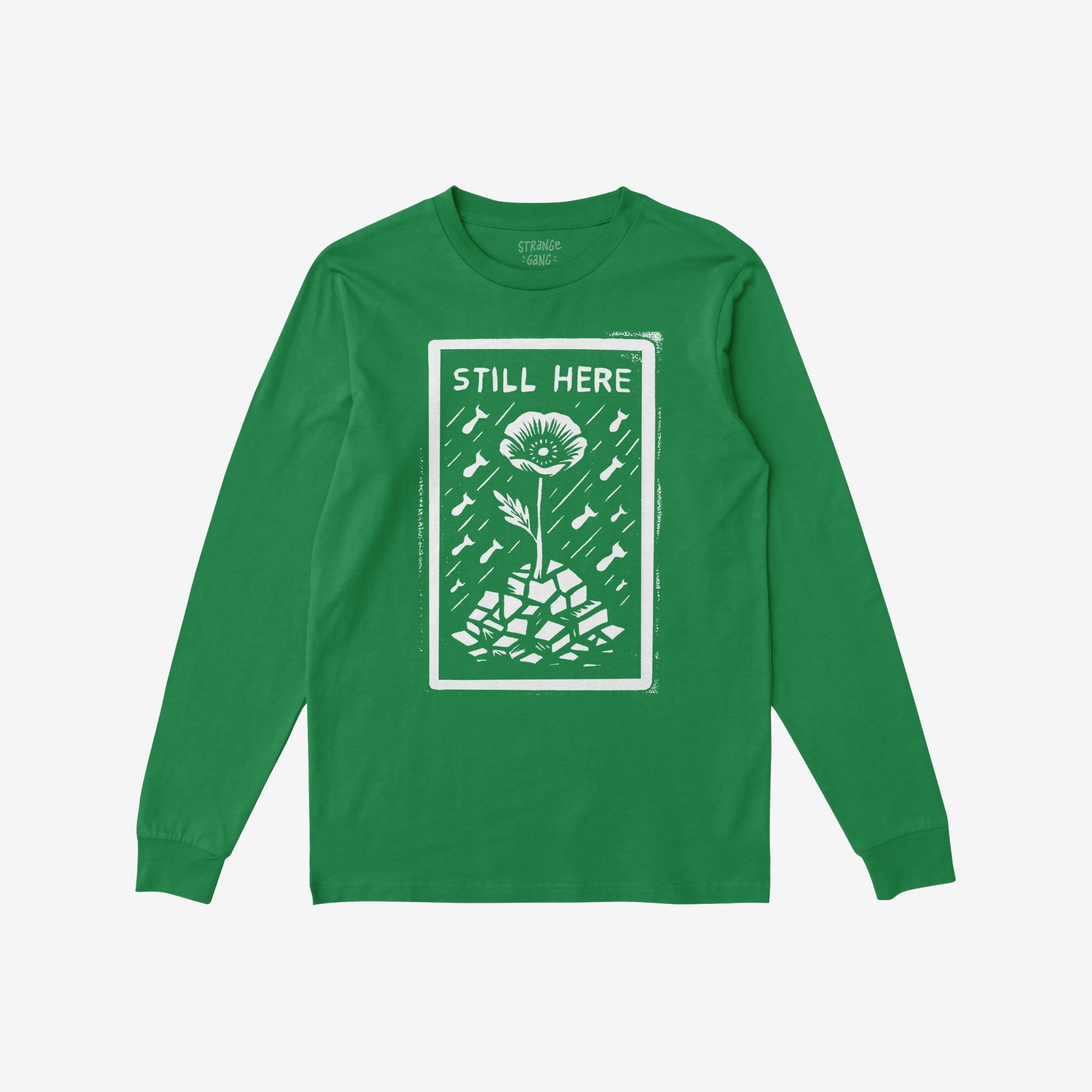 Still Here T-shirt: Green long-sleeve featuring a bold design with 'STILL HERE' text, a poppy flower emerging from rubble, surrounded by raindrops and bomb-like shapes, symbolizing resilience against oppression.
