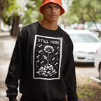 Young person in an orange bucket hat wears a Still Here JULZ Sweatshirt with a bold STILL HERE graphic, symbolizing resilience through a flower amidst falling bombs, on a tree-lined sidewalk.
