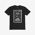 The Still Here T-shirt features a linocut-style poppy growing through rubble, surrounded by falling shapes, symbolizing resilience and protest, aligning with anti-war, pro-freedom values.