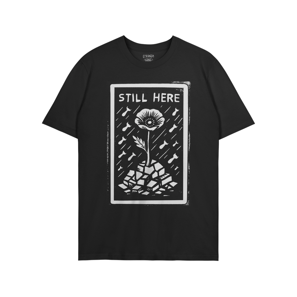 The Still Here T-shirt features a linocut-style poppy growing through rubble, surrounded by falling shapes, symbolizing resil