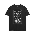 The Still Here T-shirt features a linocut-style poppy growing through rubble, surrounded by falling shapes, symbolizing resil